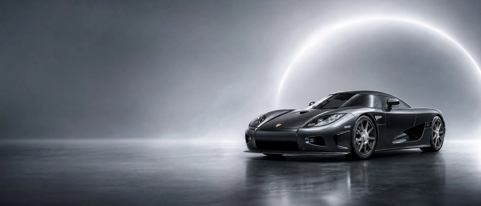 A metallic black Koenigsegg CCX, front three-quarter view on a silver-graphite field with a sun halo