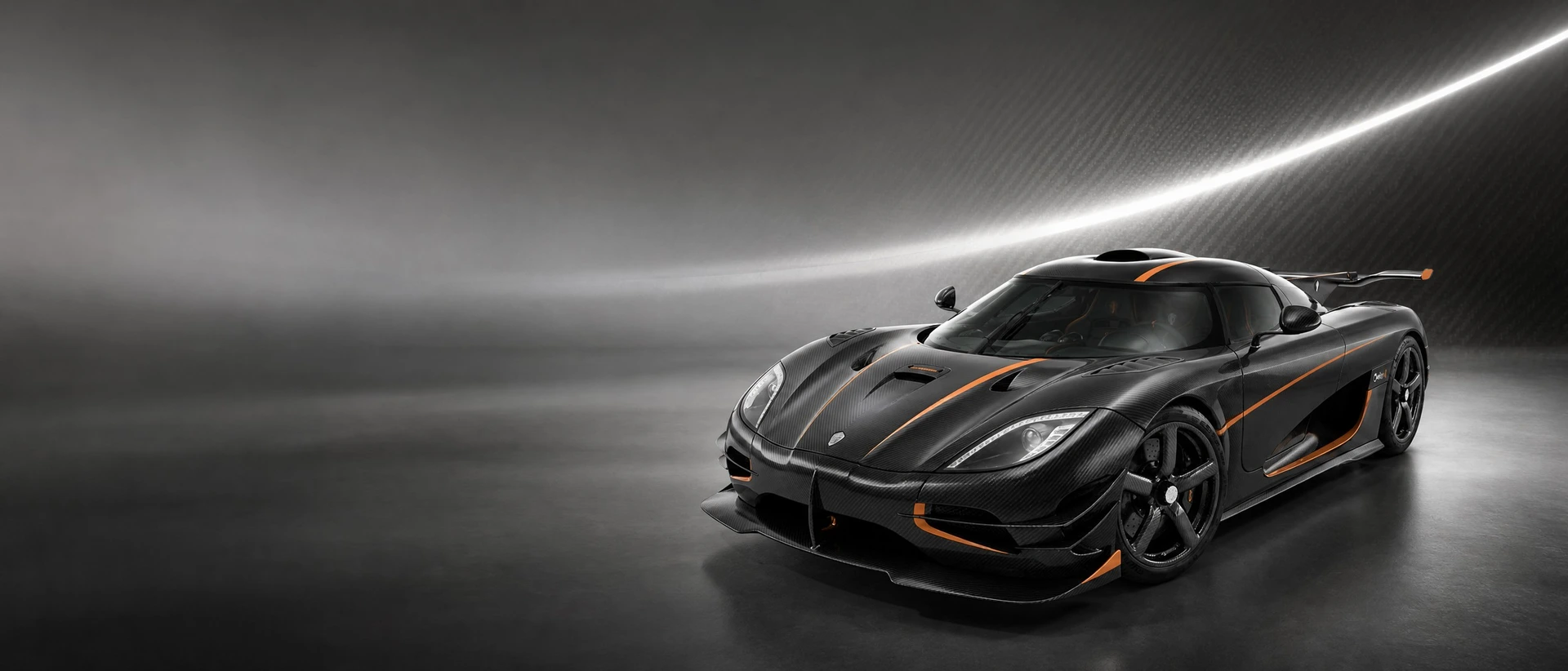A black carbon Koenigsegg One:1, front three-quarter view on a graphite field with a taut line of light