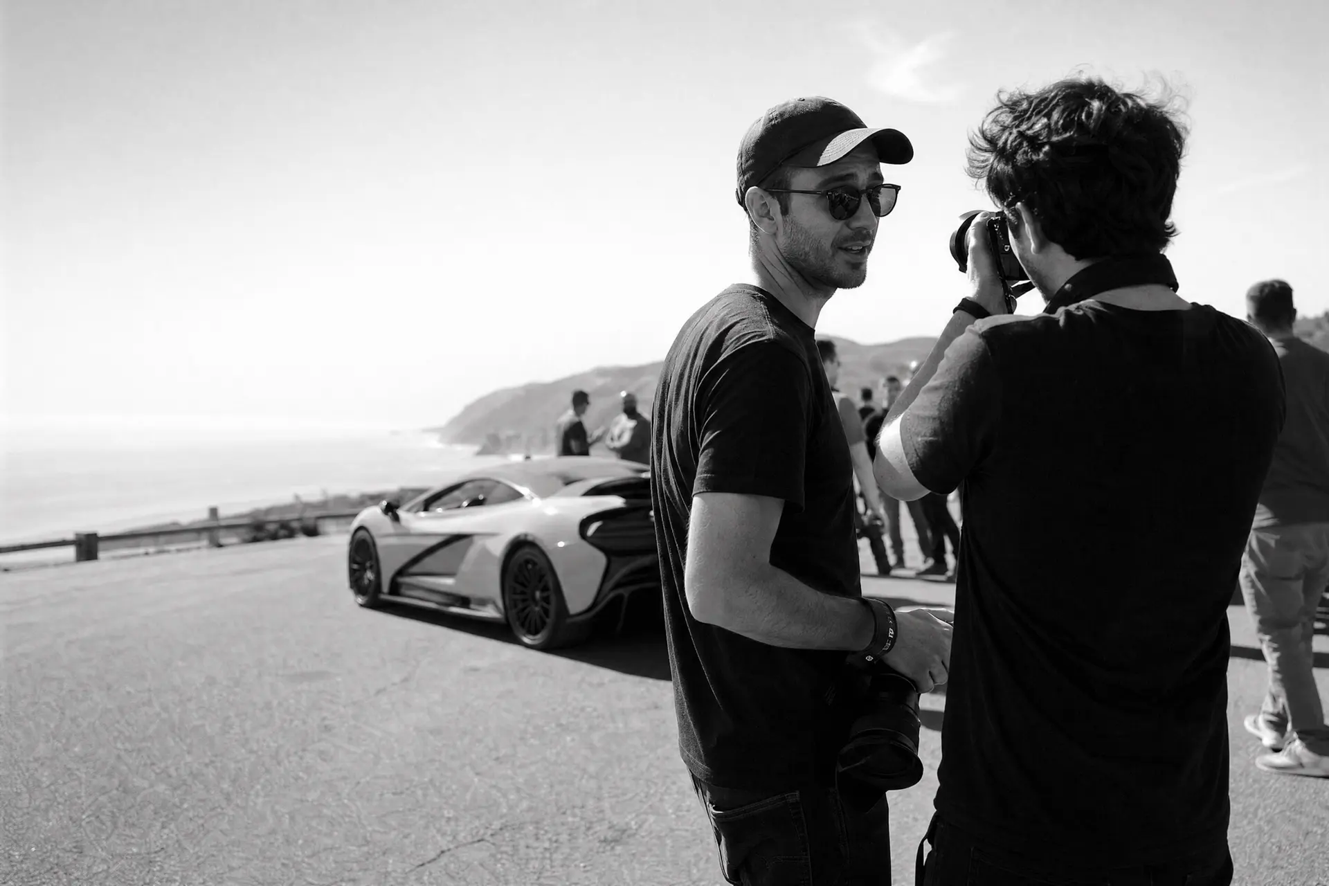Two car photographers in conversation at a coastal tour stop, one raising a camera, a modern hypercar parked behind them in daylight