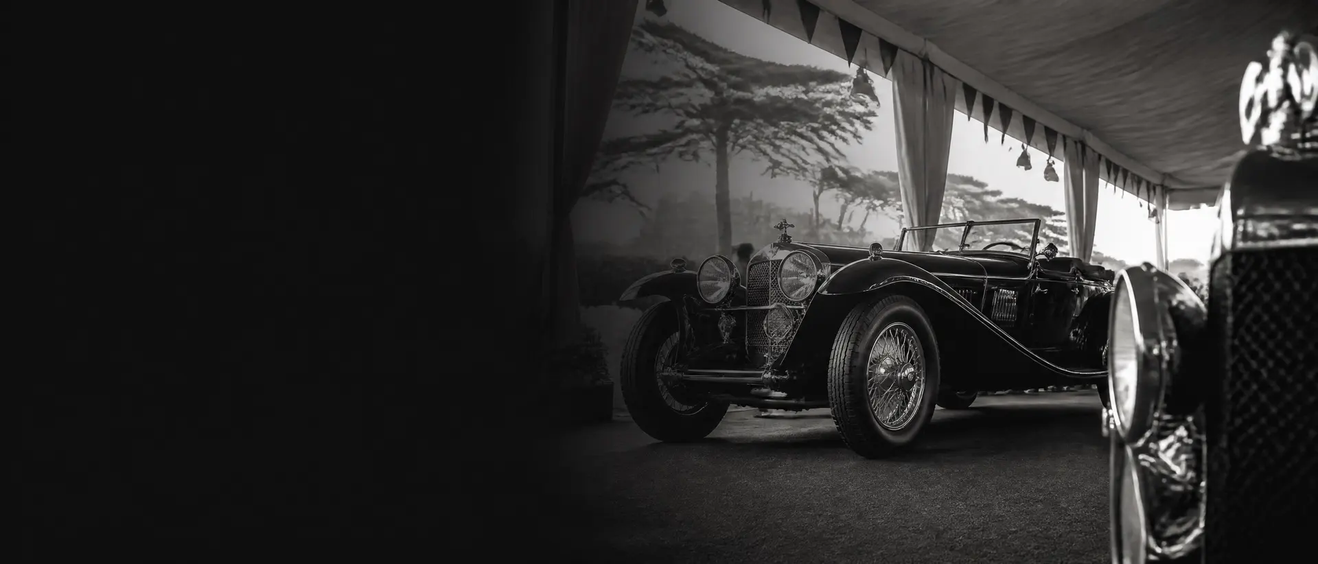 Black-and-white image of a pre-war grand tourer under an auction tent’s rigging, monochrome pennants, Monterey pines behind, a chrome radiator shell cropped in the foreground