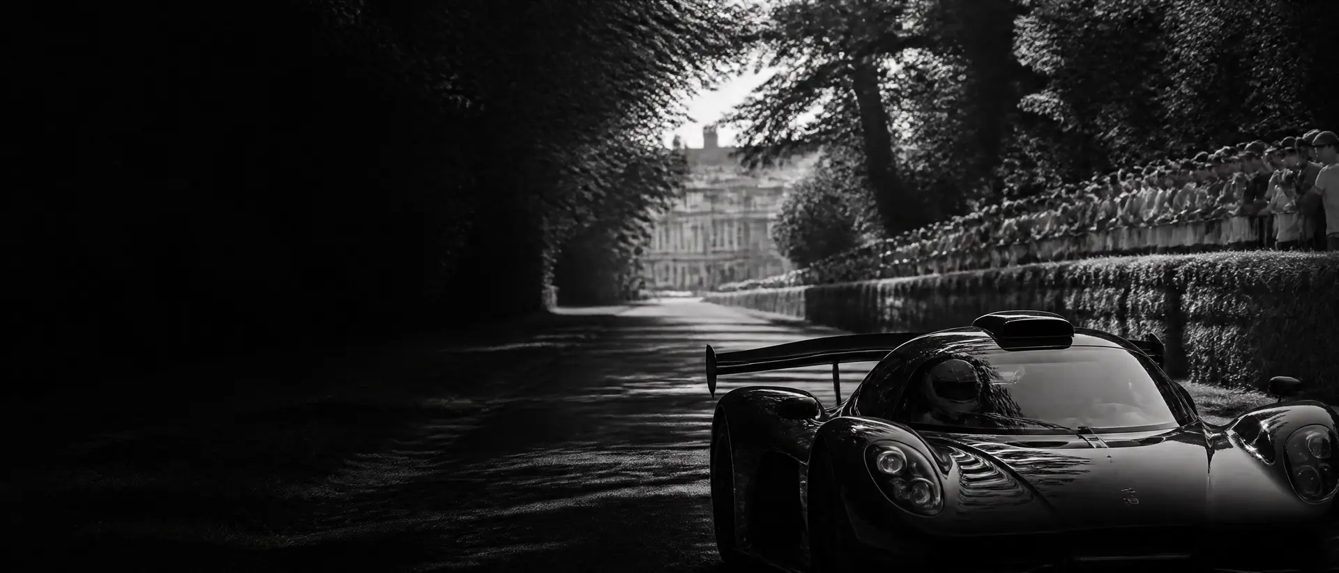 Black-and-white image of a hypercar launching off the line on a tree-lined estate driveway, hay bales and crowds along the fence, heat haze rising, the car’s splitter cropped at the lower edge