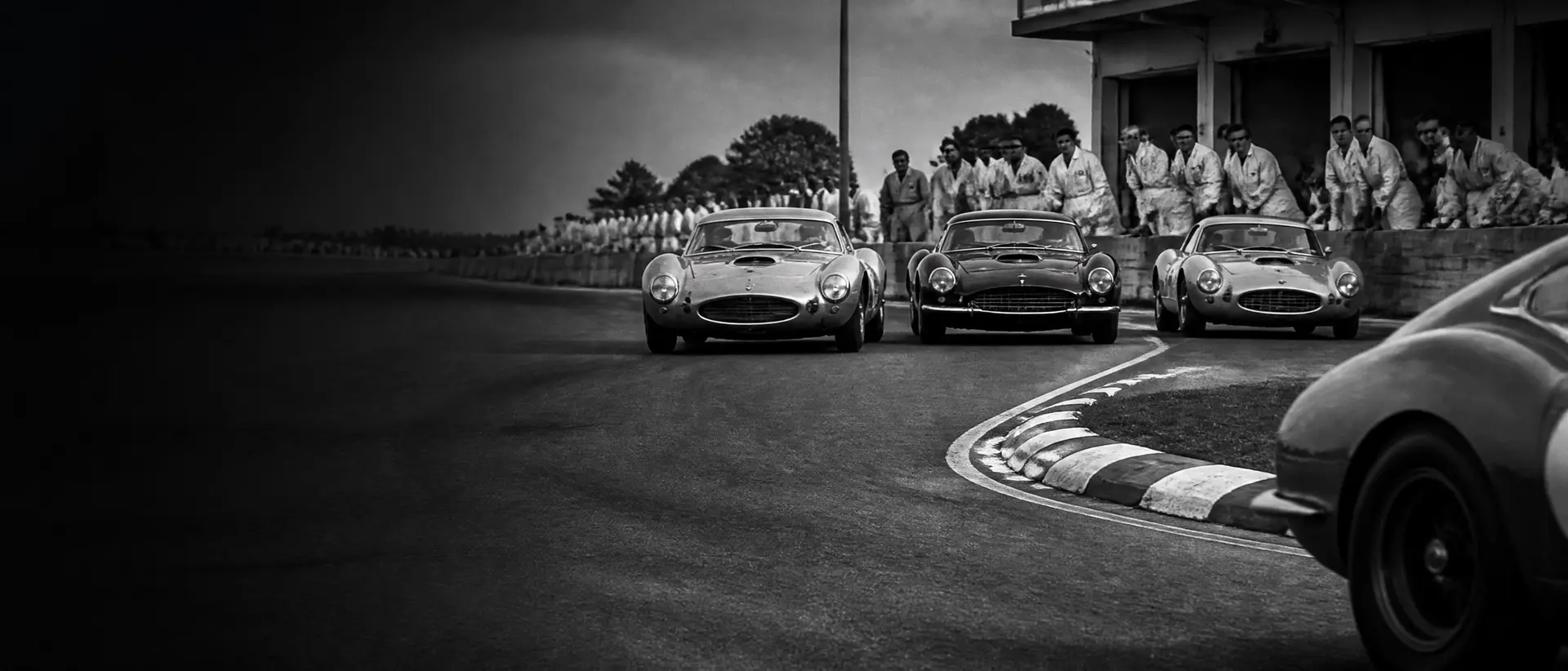 Black-and-white racing scene at a period circuit, 1960s GT cars three abreast into a corner, pit crews in period overalls, chequered kerb and a cropped rear haunch in the foreground