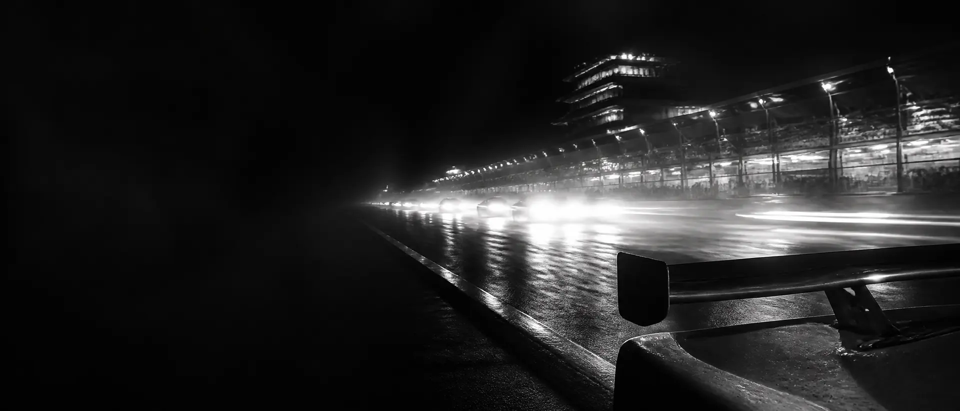 Black-and-white night scene at a race circuit, headlights streaking past the pit wall, a long-tail prototype’s rear wing cropped in the foreground, floodlit signage reduced to abstract shapes