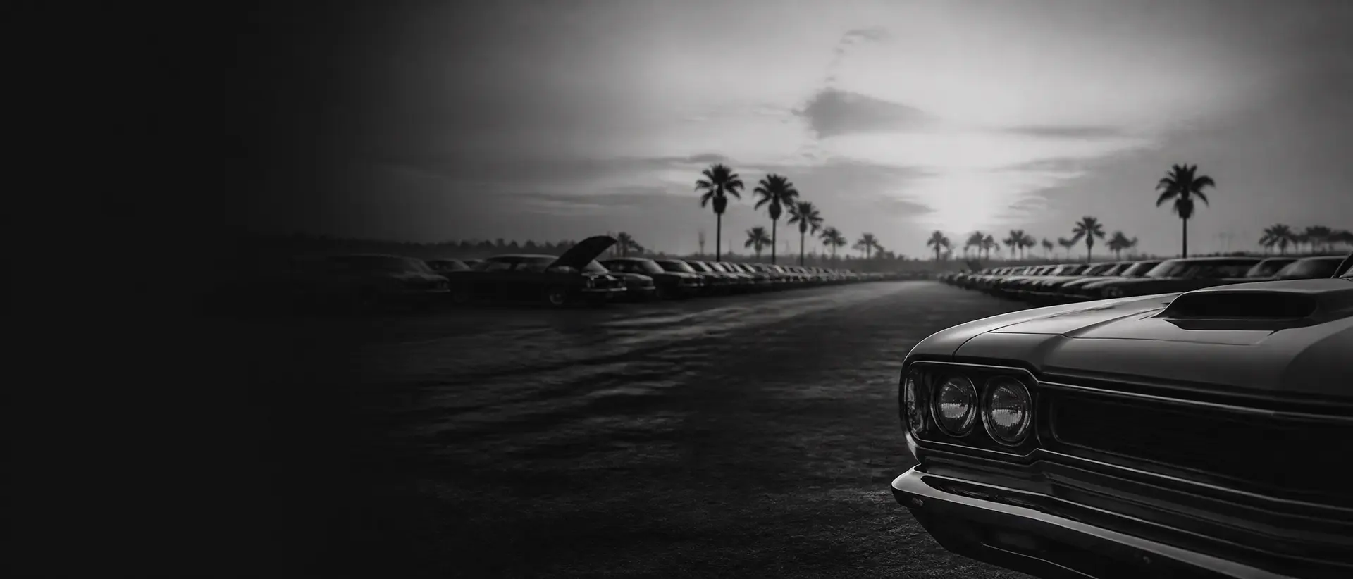Black-and-white panorama of an endless auction paddock at dawn, rows of collector cars receding into Florida haze, the nose of a muscle car cropped hard at the frame edge