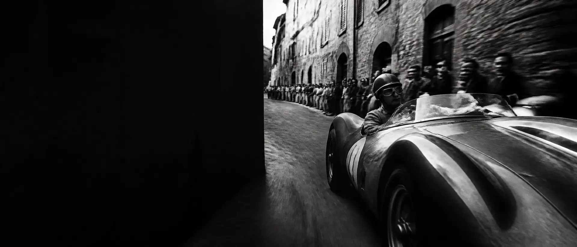 Black-and-white action shot of a 1950s open sports racer sweeping through an Italian hill town at speed, stone walls blurred, crowd leaning in, the bonnet cropped by the frame edge