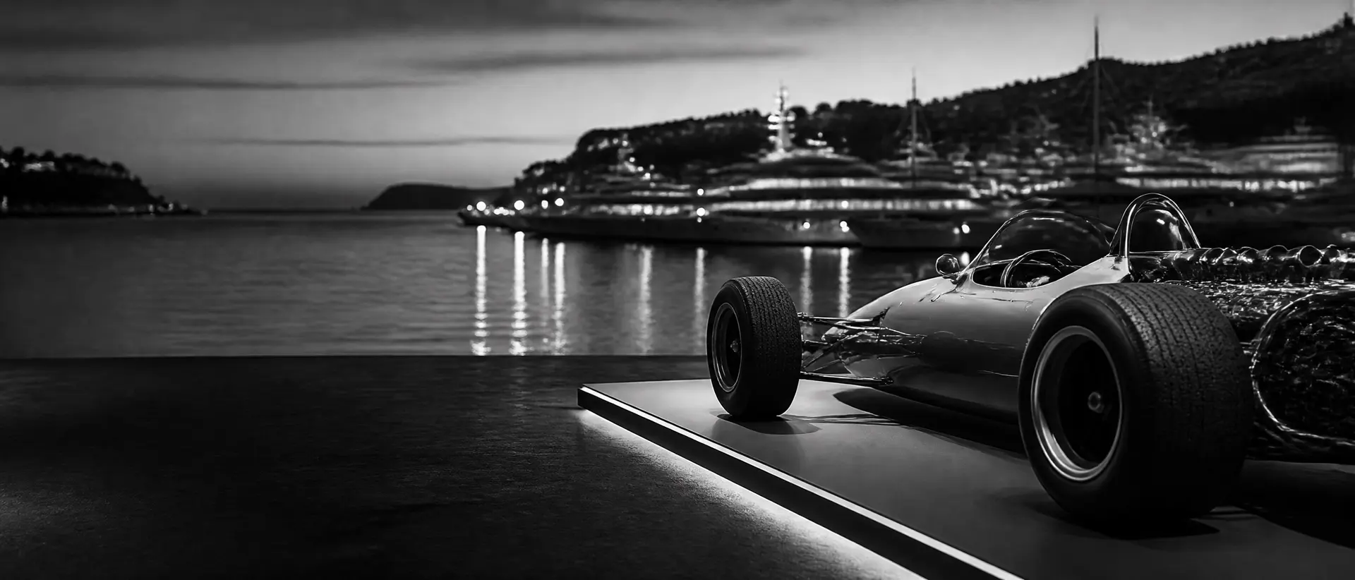 Black-and-white scene of a 1960s Formula car on a plinth against Monaco harbour, superyachts soft in the background, the car’s exposed engine cropped at the frame’s right edge