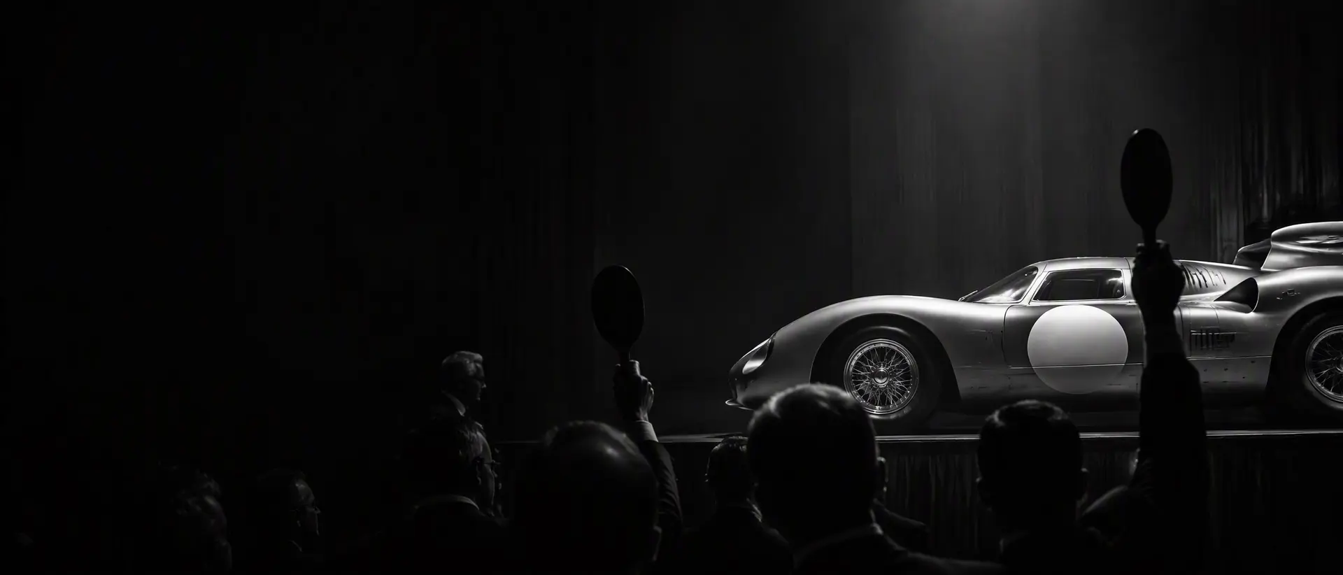 Black-and-white auction room scene, a 1960s Ferrari prototype on the block under a single spotlight, paddles raised in silhouette, the car’s tail section cropped by the frame