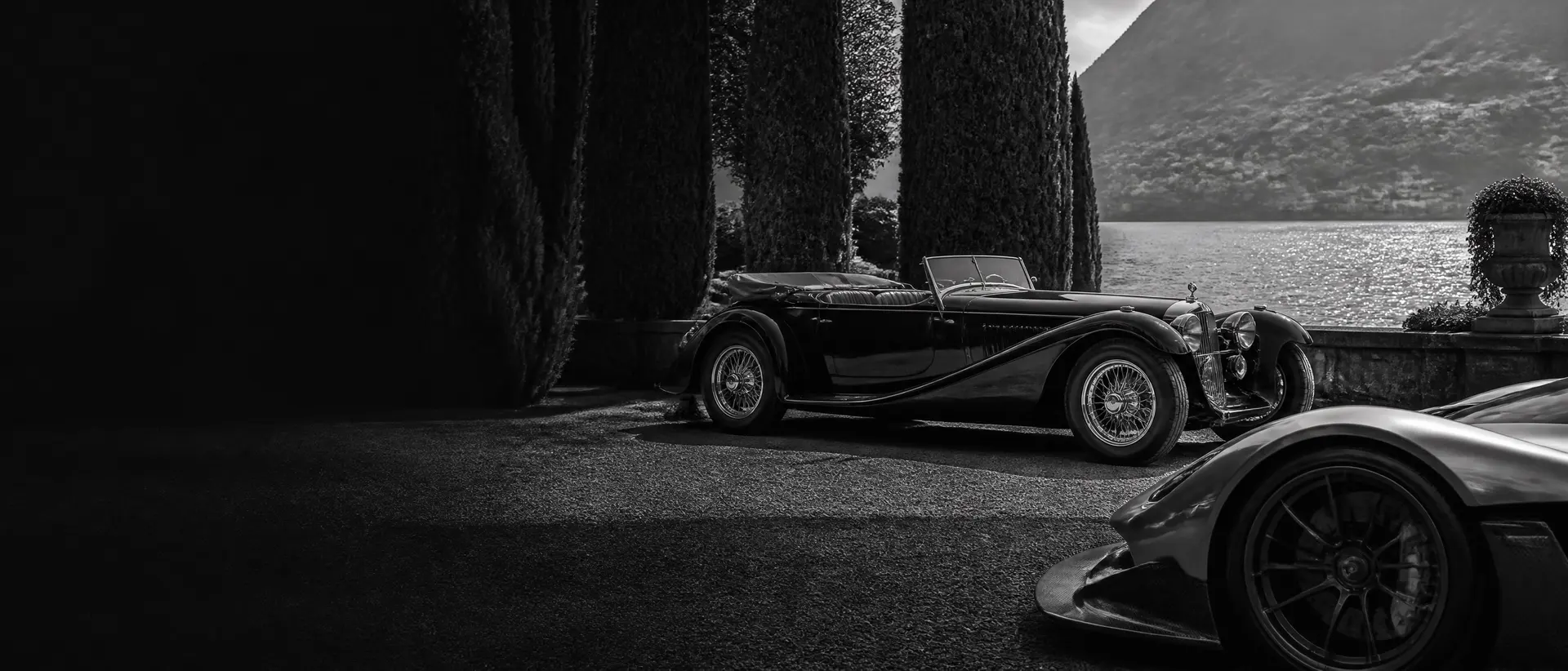 Black-and-white composition of a 1930s coachbuilt cabriolet under cypress trees, Lake Como glittering monochrome behind, a hypercar’s front wing cropped in the near foreground