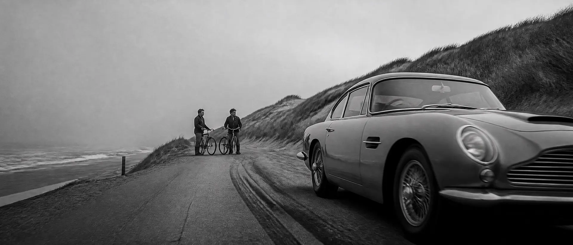 Black-and-white scene of a 1960s GT driving a beach road between dunes, North Sea mist, cyclists paused to watch, the car’s long bonnet cropped by the right edge of the frame