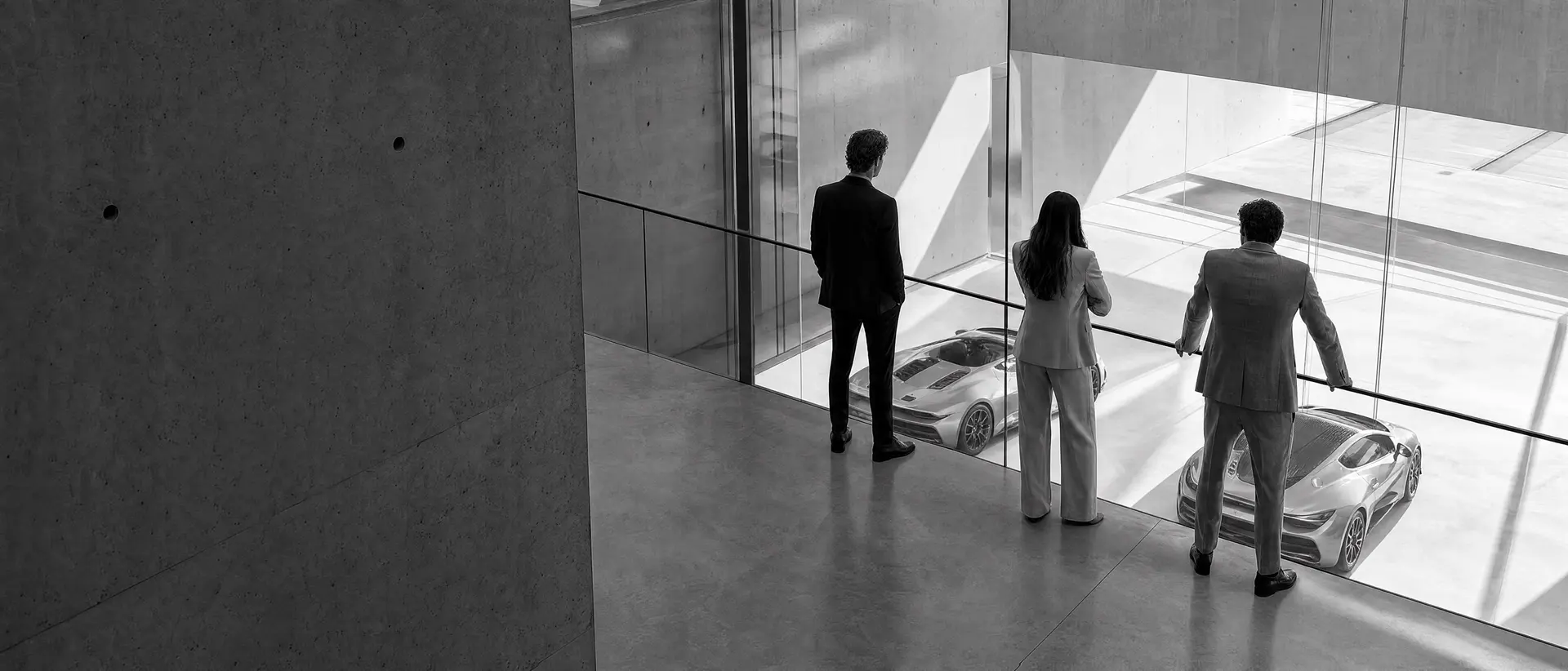 Three people at a glass rail on the mezzanine of a modern collection building, looking down on two cars in daylight