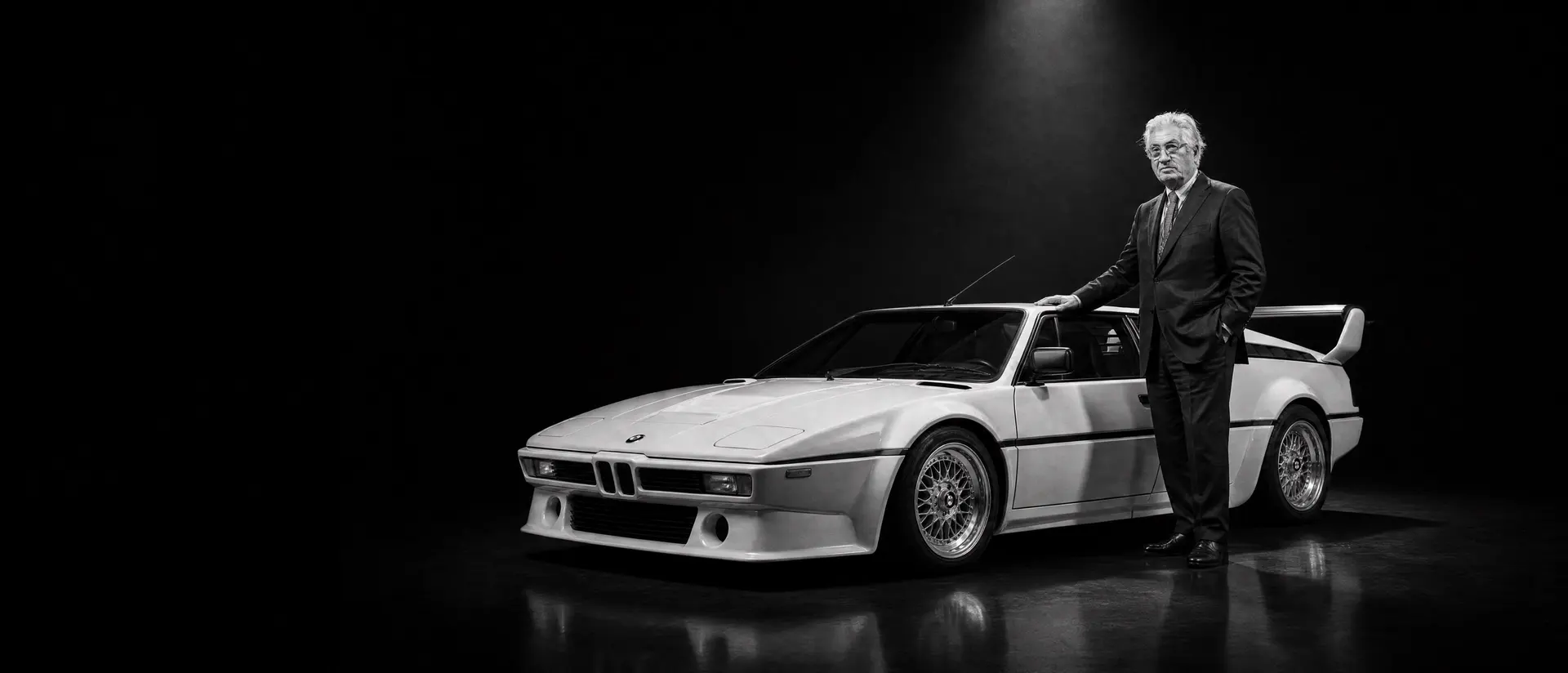 Giorgetto Giugiaro standing beside a BMW M1 under a single spotlight